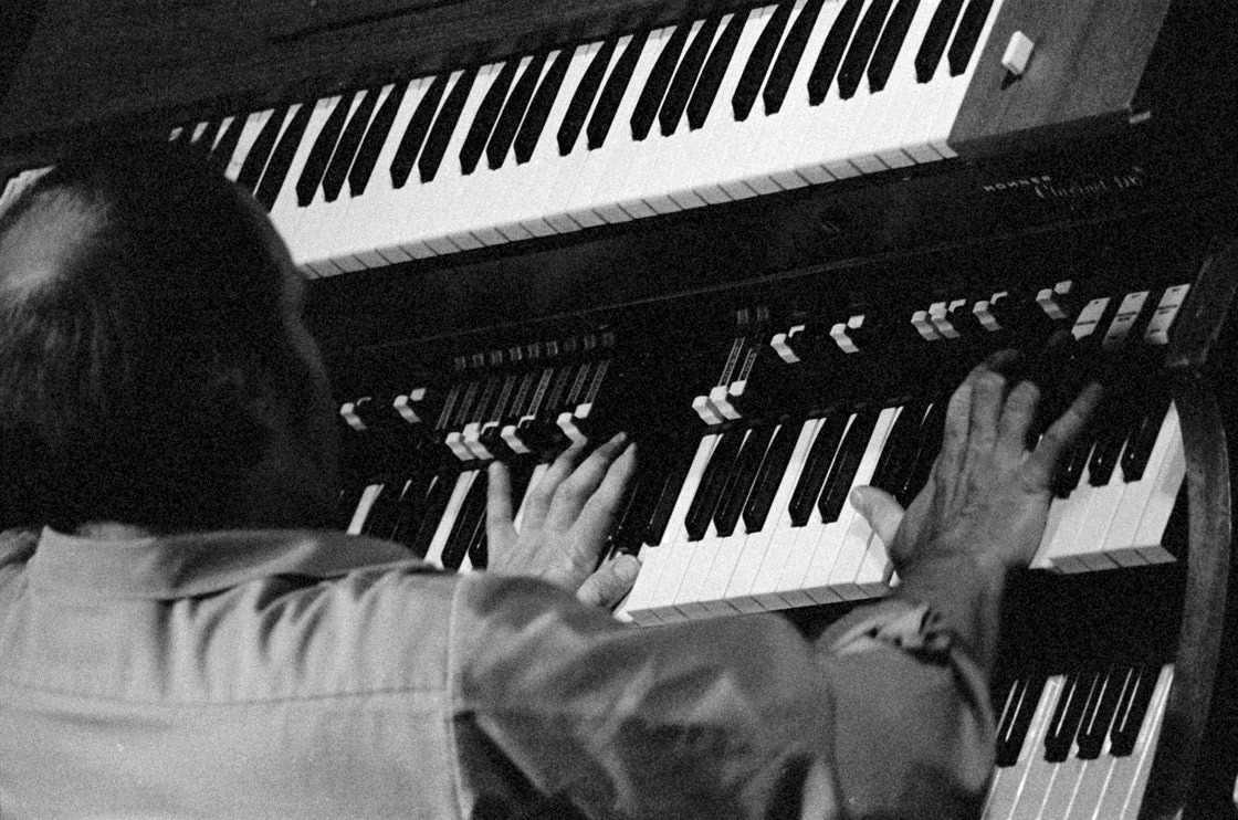 Close up from over the shoulder, two hands spread across the lower manual of an organ in black and white.